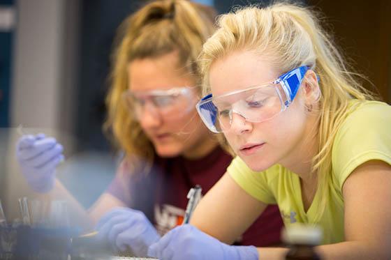 Photo of two female Chatham University students wearing protective goggles and writing at a lab table