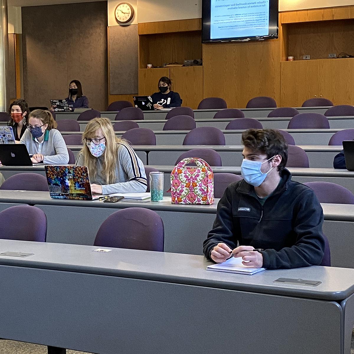 Photo of masked students seated at a distance from one another in a lecture hall, working in notebooks and on laptops