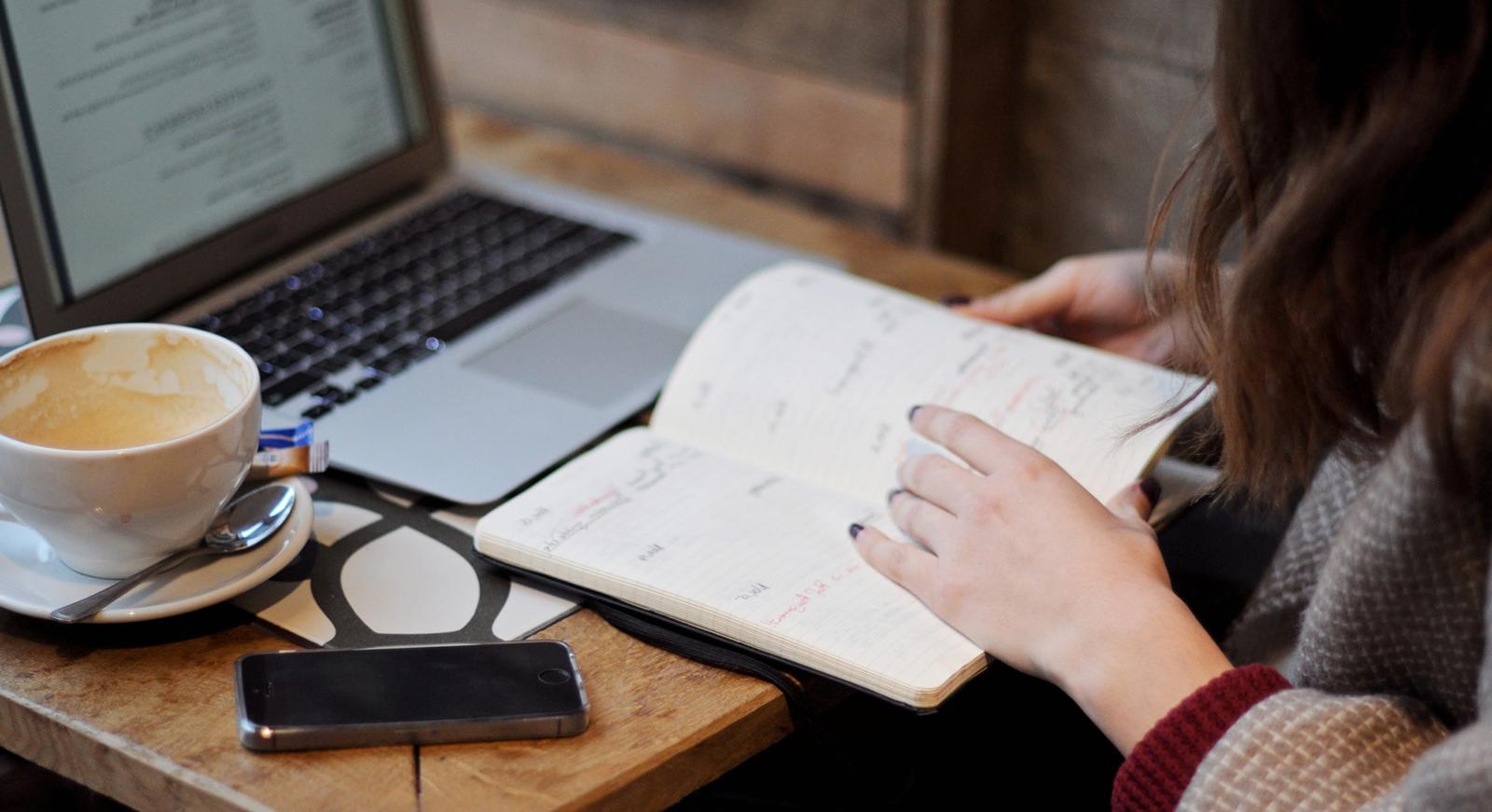 Close-up photo of a woman sitting in a cafe with coffee, a laptop, and a notebook on the table in front of her.