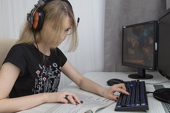 Photo of a Chatham University student working on computer while wearing headphones and referencing a textbook. 