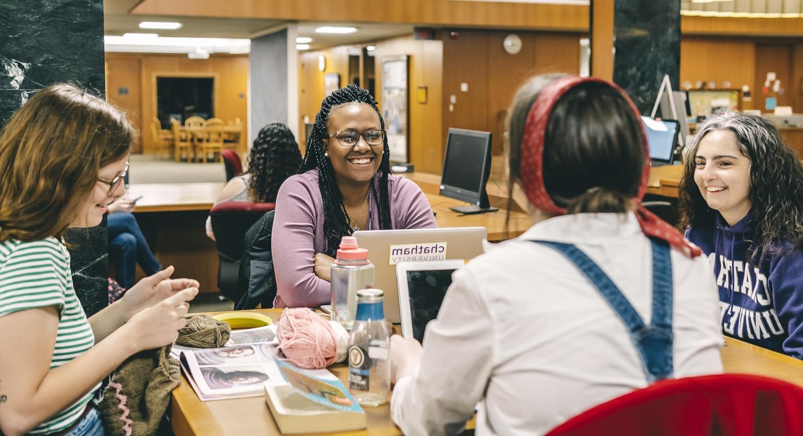 Photo of four female Chatham University students sitting at a table in Jennie Mellon King Library talking with laptops, notebooks, and knitting. 