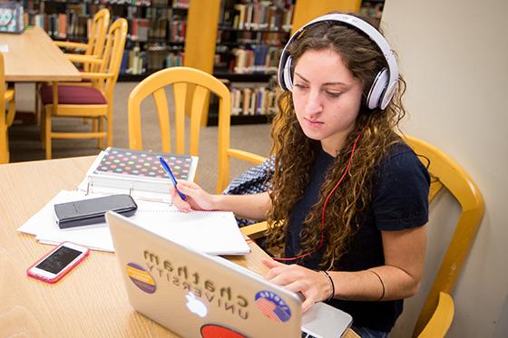 Photo of a Chatham University student sitting at a table in Jennie Mellon King Library studying on a laptop and writing in a notebook with a calculator. 