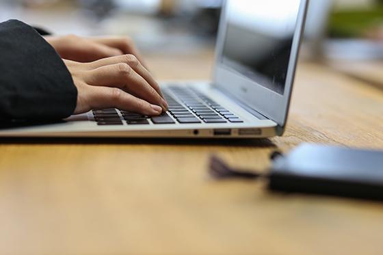 Close-up of a photo typing on a laptop, with a notebook on the table