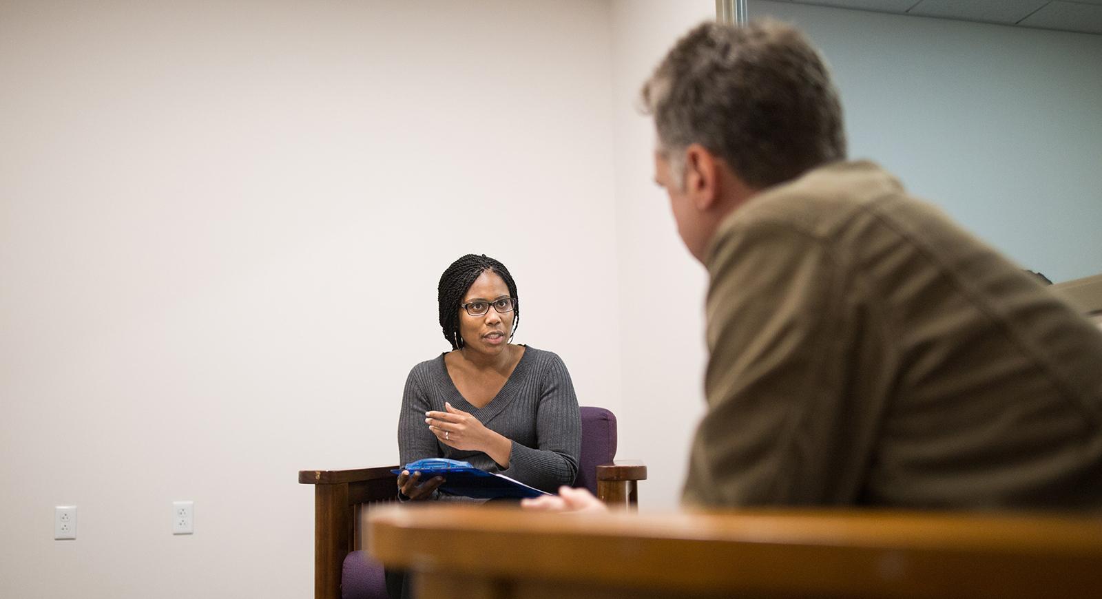 Photo of a man and a woman sitting opposite of one another in chairs while talking in a clinical setting. 