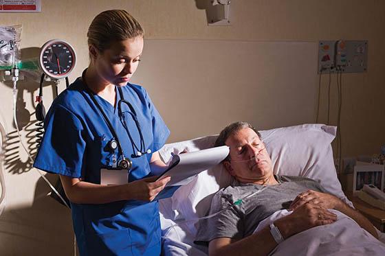 Photo of a nurse in scrubs, writing on a clipboard next to a sleeping patient